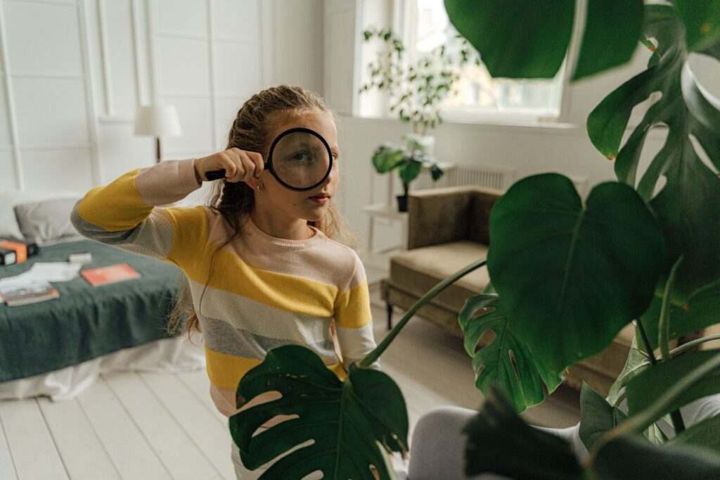 Young girl using magnifying glass to explore indoors surrounded by green plants.