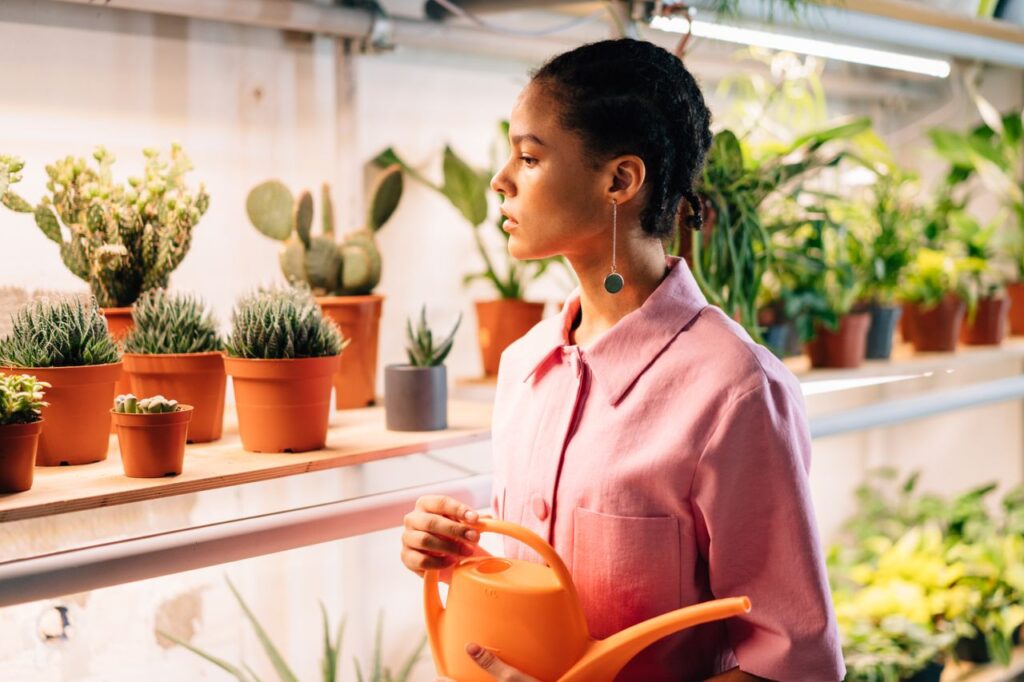 A woman in a pink shirt waters indoor potted plants in a modern studio.